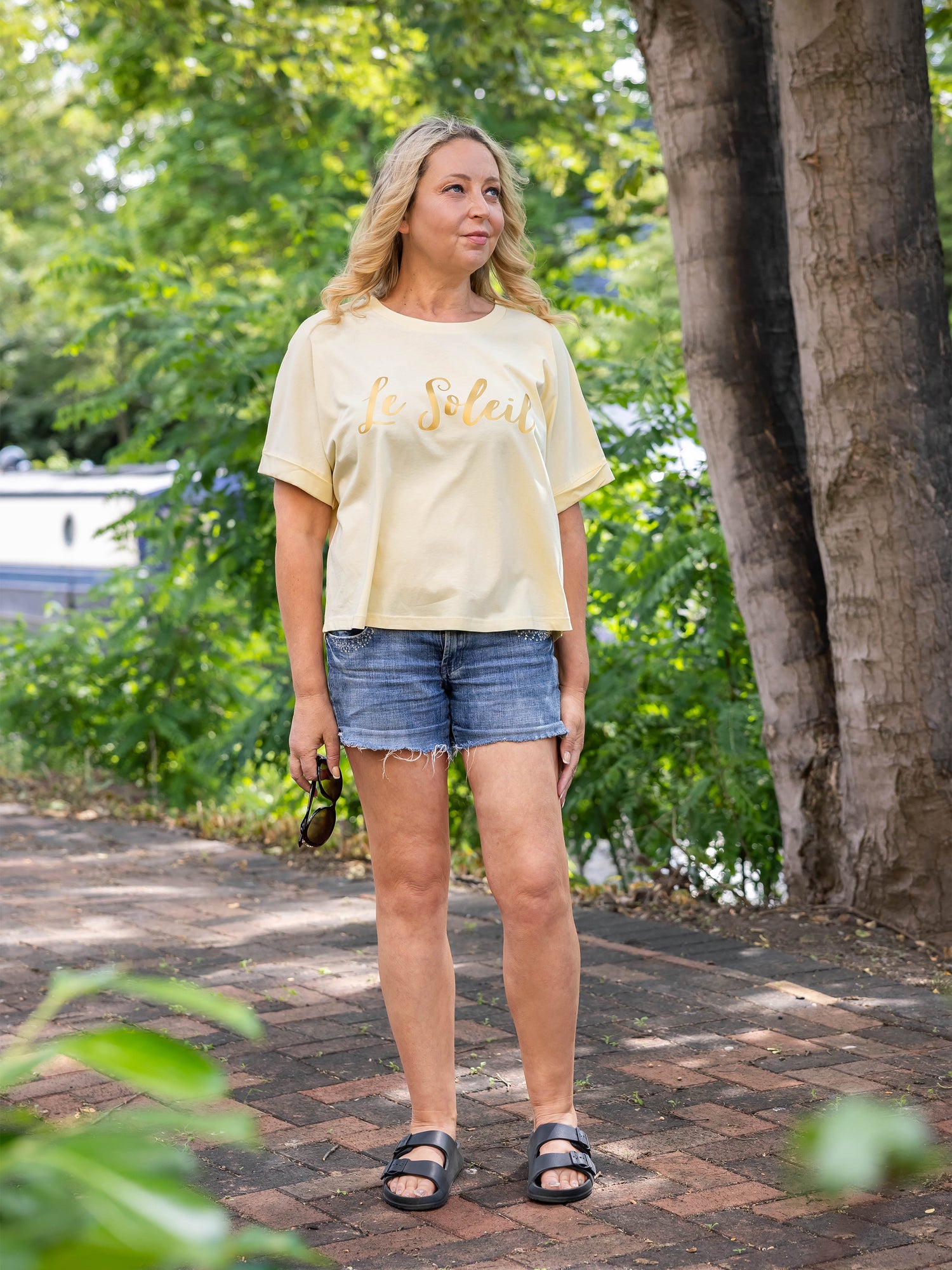 Woman wearing a light yellow t-shirt with text and denim shorts standing. Woman sitting on a wooden weathered chest. Butter light yellow oversized summer t-shirt with loose short sleeves and rolled cuff. Round neck Ladies t shirt with slogan design Le Soleil in matt metallic gold script font.