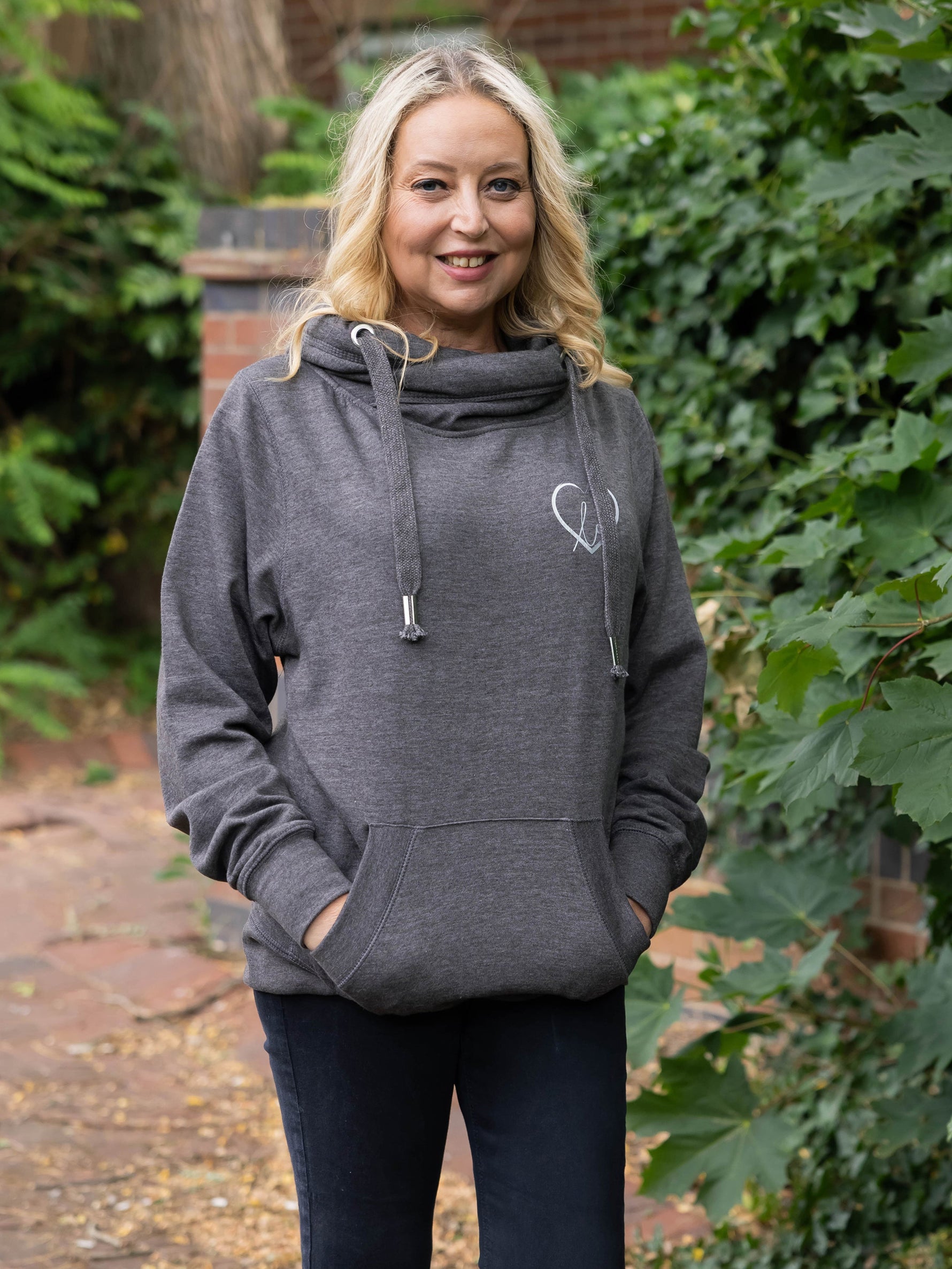 Model outdoors in nature wears womens Cowl Neck Hoodie - Dark Grey hoodie with silver metallic love heart motif. Charcoal ladies hoodie with Long sleeves and thumb loops to cuffs. Front kangaroo pouch pocket. Cross neck sweatshirt with hood