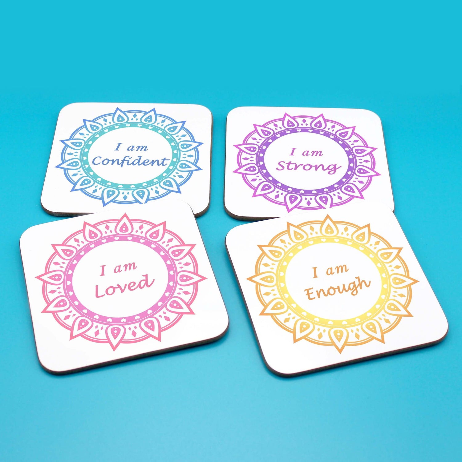 Wellbeing gift. Four square wooden backed positive affirmations coaster set. Mandala design with daily affirmation wording inside the mandala. Affirmations read I am Loved (red and pink mandala), I am Enough (yellow and orange mandala), I am Confident (blue mandala), I am Strong (Purple mandala)