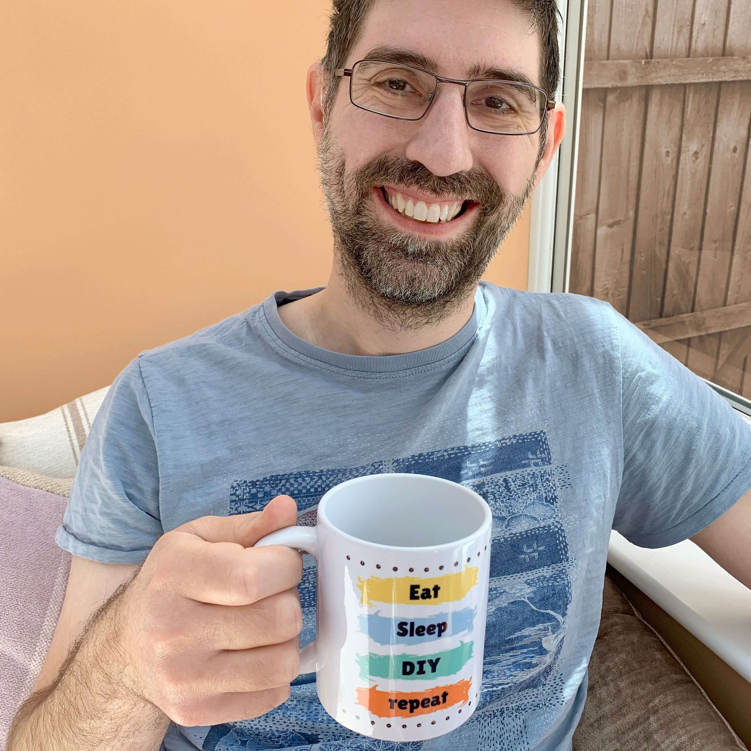 Dad gifts idea. Man smiling wearing a blue t-shirt with short sleeves. Holding a white ceramic mug with line of screws top and bottom and 4 different colour paint stripes design. Each stripe of paint has a word in vertical sequence. Eat Sleep DIY repeat.
