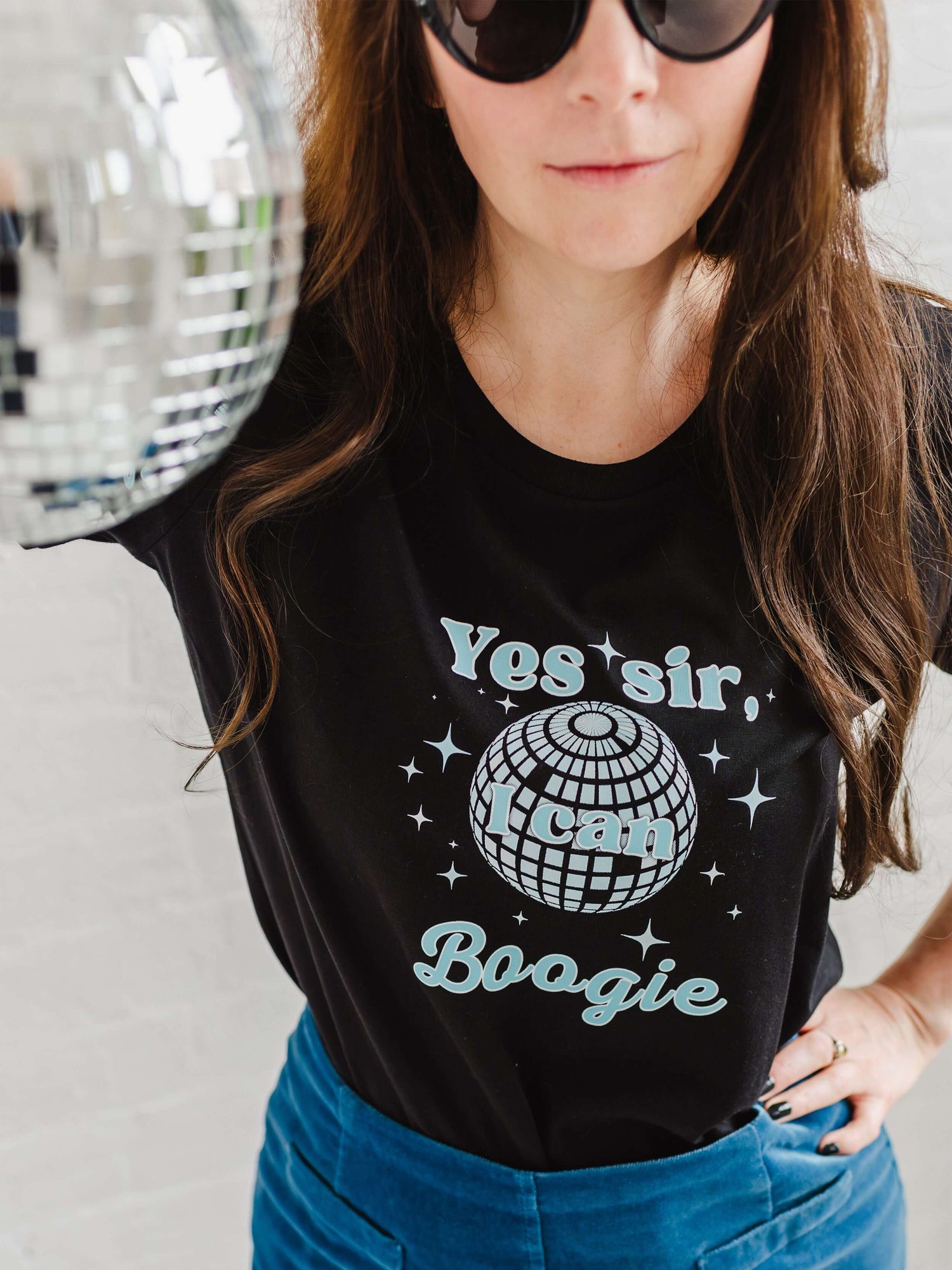 Close up of Ladies Black graphic t-shirt with mirror ball design. Woman holding a silver disco ball, worn with a velvet A line mid blue skirt. Fitted short sleeve tshirt with round neck. 70s disco music inspired ladies slogan tshirt with disco ball design to background of top and sparkles surround. Slogan text in foreground reads Yes sir I can Boogie in light blue font. Organic cotton casual fitted Tee
