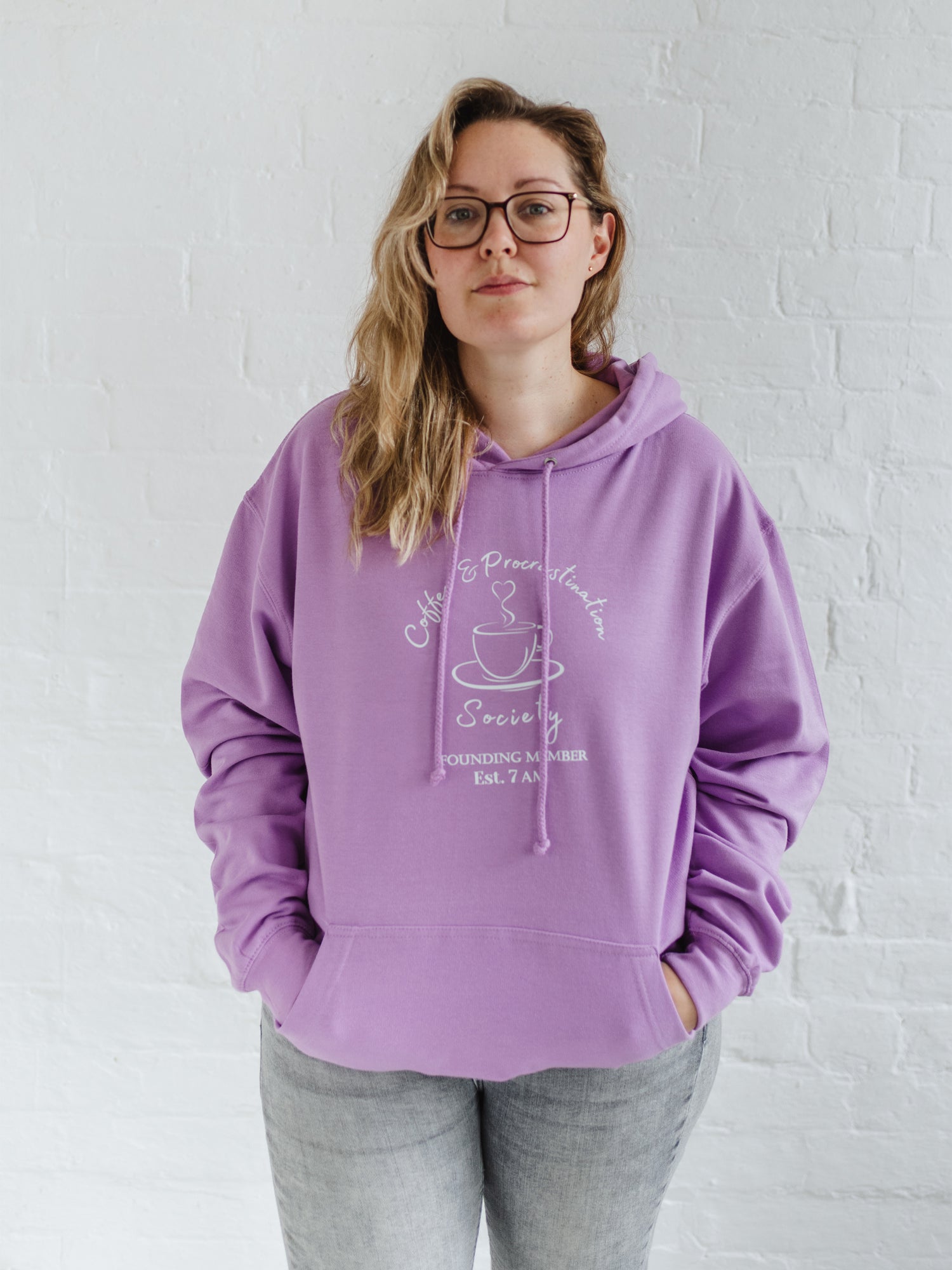 Woman with blonde long hair standing wears a womens coffee themed oversized fit hoodie. Lavender purple hoodie top with coffee cup and saucer design. White outline with steam from cup shaped into a heart. Writing above reads Coffee & Procrastination. Below reads Society FOUNDING MEMBER Est. 7am. She pairs it with grey skinny jeans
