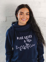 Womens Navy blue hoodie with cross cowl neck. Thumb holes to sleeves. Design is of four light grey butteflies in flight and wording Blue Skies and Butterflies to the front. Unisex relaxed fit hoodie. Light purple wording with light grey back shadow. Long sleeves and straps from cowl neck with metal toggles on the end. Worn by caucasian woman with long dark brown hair smiling