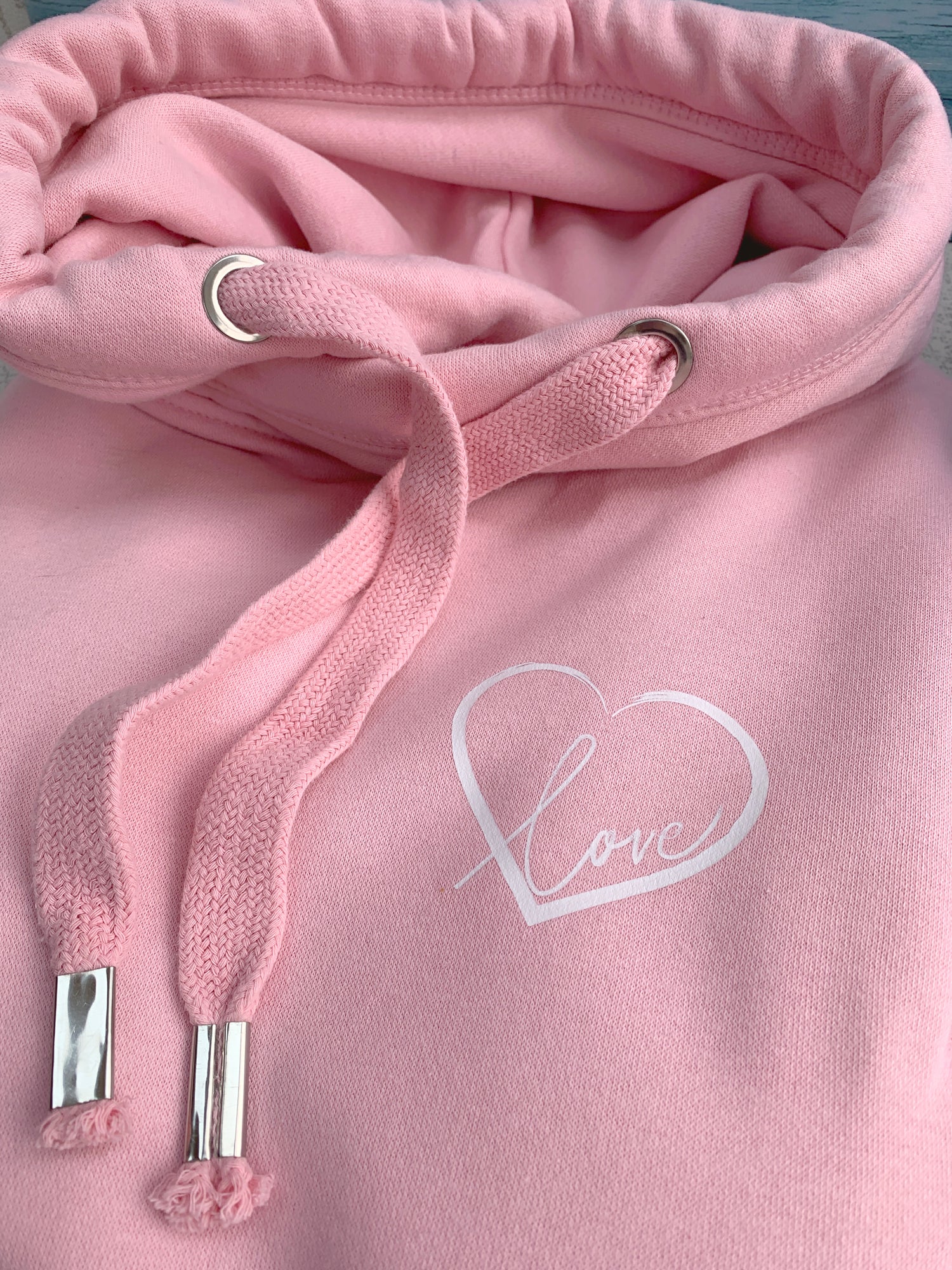 Close up to baby pink womens hoodie Love heart cross cowl neck casual fashion hoodie. Cotton Polyester blend relaxed fit unisex oversized style hoodie design has love heart. Whitert to upper left of the chest with word Love in white script font nestled within the heart.