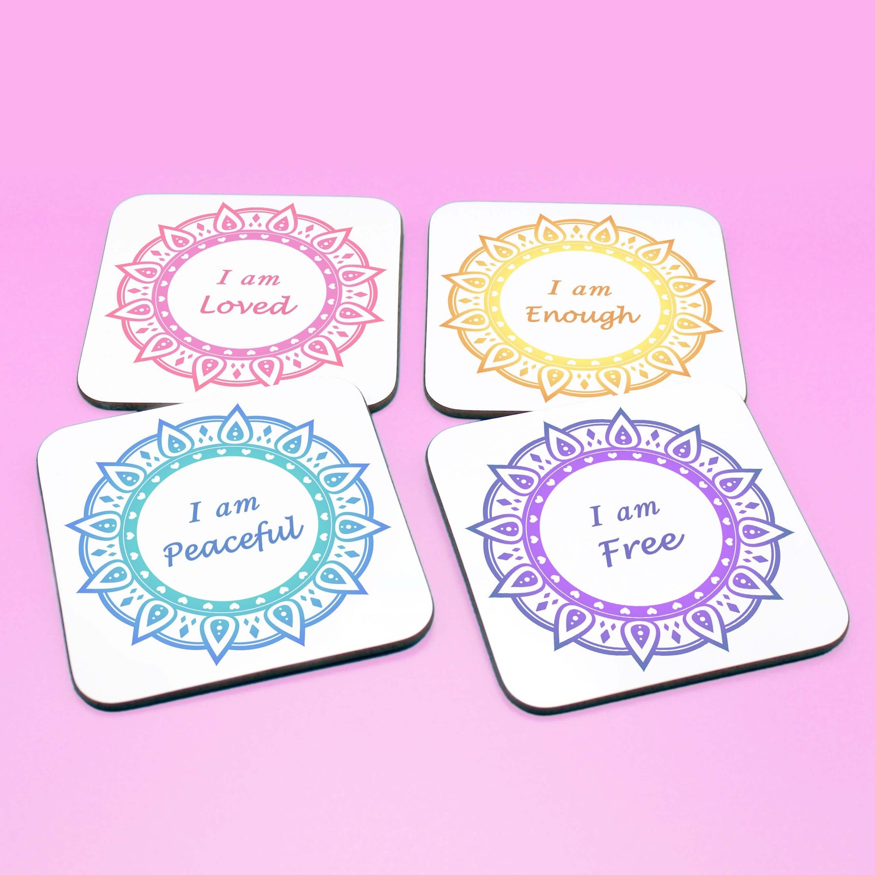 Wellbeing gift. Four square wooden backed personal affirmations coaster set. Theme is Letting Go in this set of four. Mandala design with daily affirmation wording inside the mandala. Affirmations read I am Loved (red and pink mandala), I am Enough (yellow and orange mandala), I am Peaceful (blue mandala), I am Free (Purple mandala)