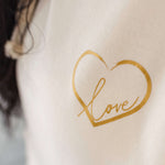 Close up of Ladies Cowl Neck Hoodie - Vanilla Cream hoodie with gold metallic love heart motif. and words Love in script font inside heart. Cross neck hood and metallic eyelet for thick cotton drawstrings with metal toggle