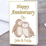 White card with gold border. Gloss A5 Happy Anniversary Greetings Card. Owl and heart design. two hand-drawn fluffy owls snuggling under interlocking gold hearts. Personalised with recipients name(s) in gold text below. Happy Anniversary wording above interlocking hearts in Gold font. Wedding Anniversary Card, white envelope