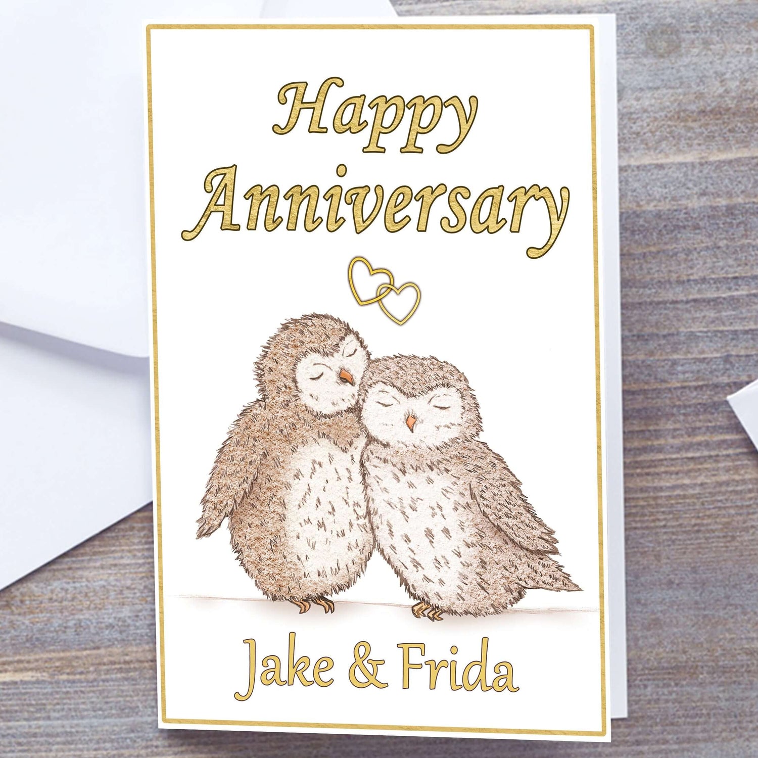 White card with gold border. Gloss A5 Happy Anniversary Greetings Card. Owl and heart design. two hand-drawn fluffy owls snuggling under interlocking gold hearts. Personalised with recipients name(s) in gold text below. Happy Anniversary wording above interlocking hearts in Gold font. Wedding Anniversary Card, white envelope