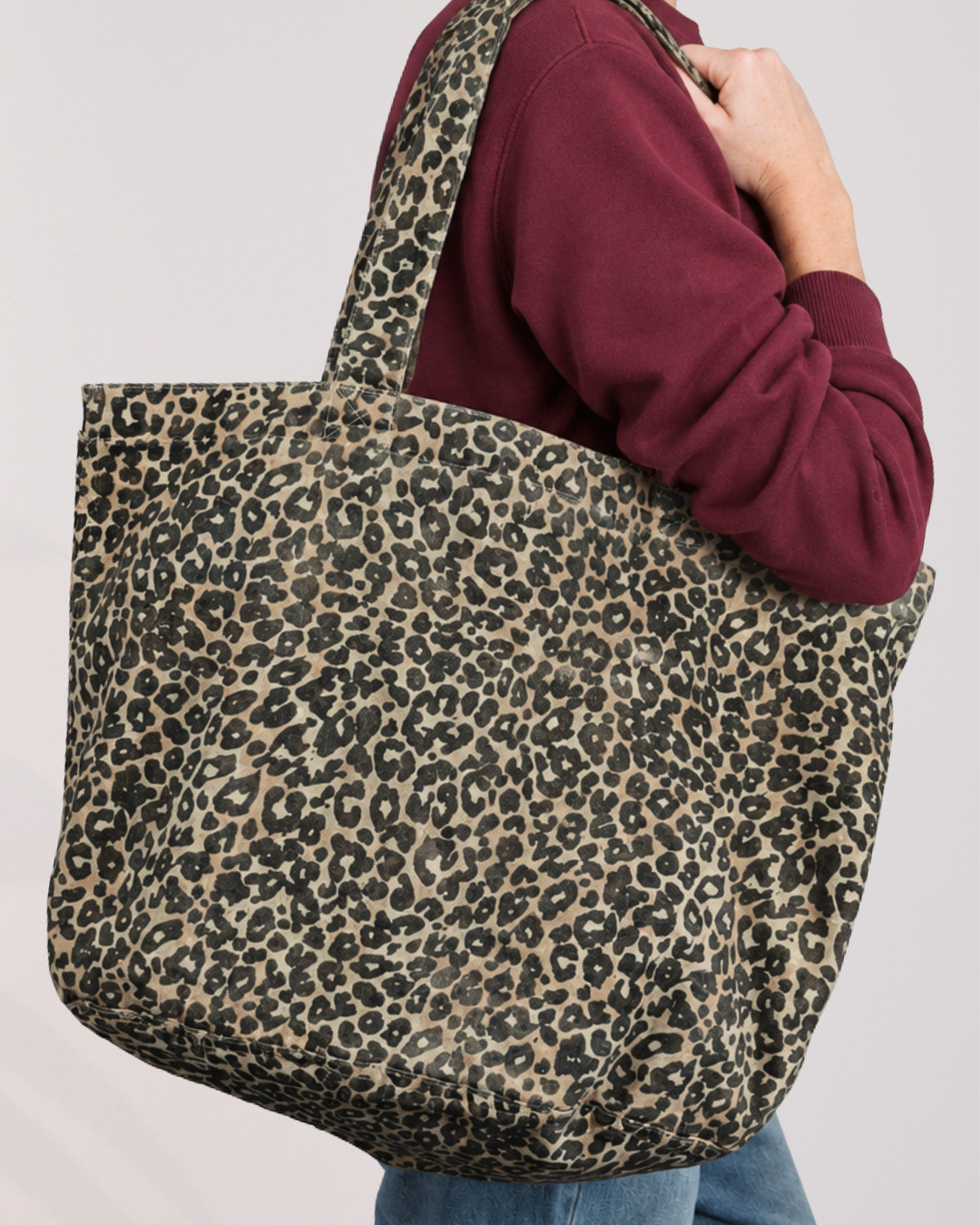 A woman wearing a burgundy sweatshirt and blue jeans carries a large canvas tote bag with a washed effect beige, brown, and black leopard print pattern against a plain light background.
