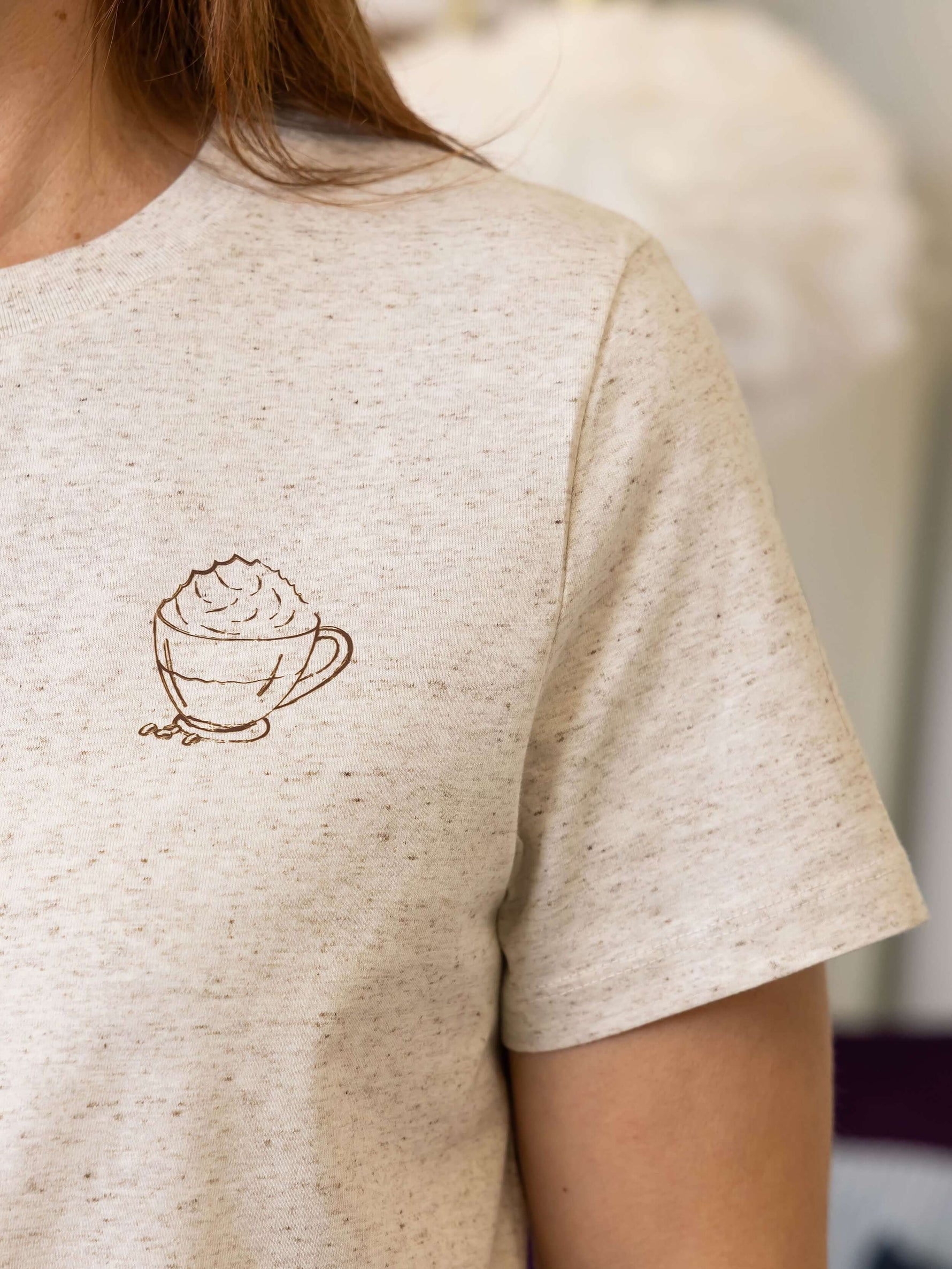 Close up of off white ladies t-shirt with a cup of coffee design to the chest. Short sleeves. Brown heather specks within organic  cotton t-shirt fabric 