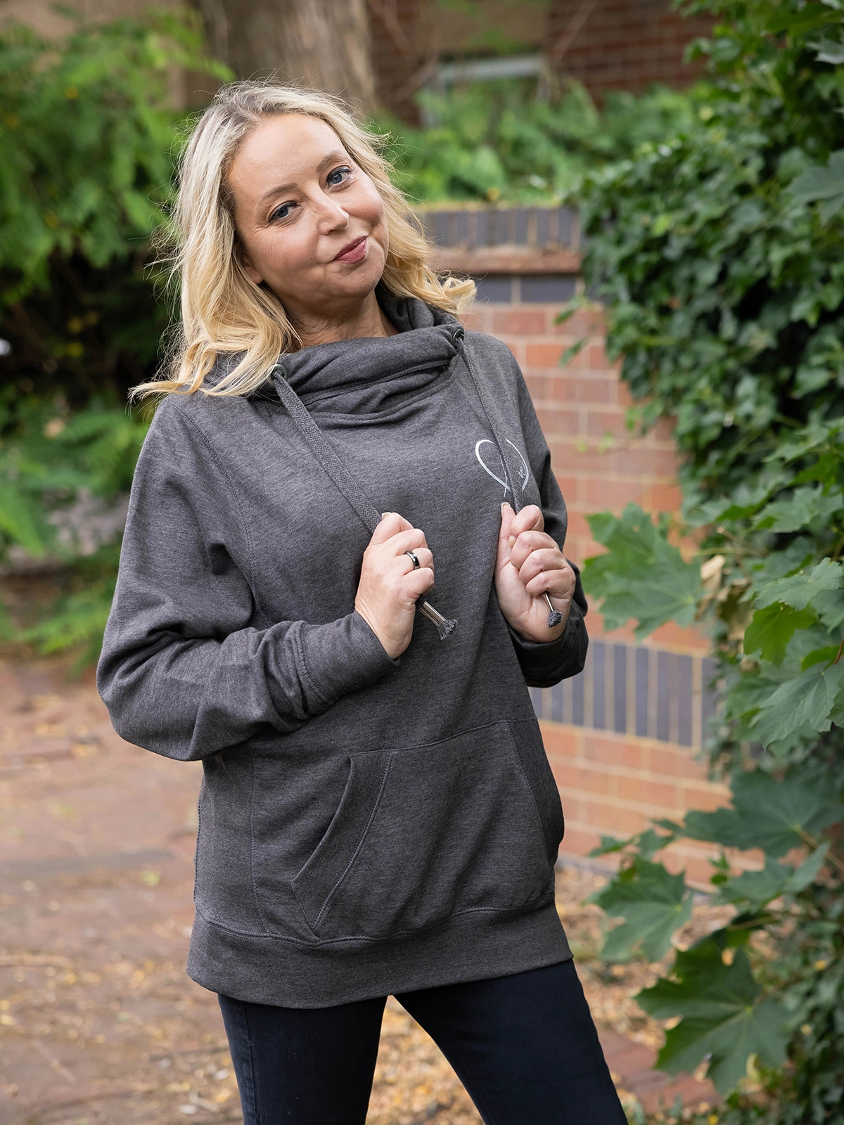 Model outdoors in nature wears womens Cowl Neck Hoodie - Dark Grey hoodie with silver metallic love heart motif. Charcoal ladies hoodie with Long sleeves and thumb loops to cuffs. Front kangaroo pouch pocket. Cross neck sweatshirt with hood