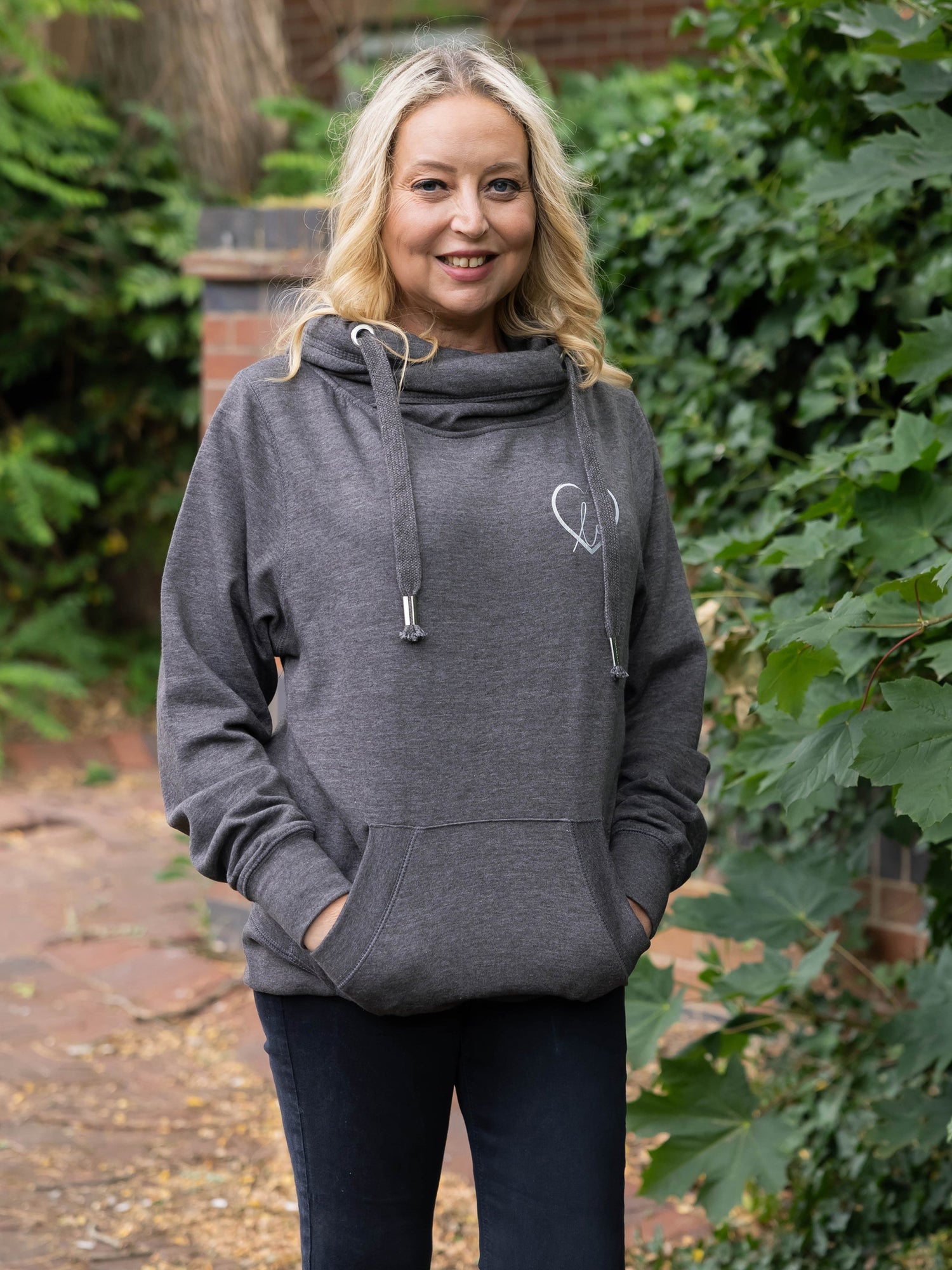 Model outdoors in nature wears womens Cowl Neck Hoodie - Dark Grey hoodie with silver metallic love heart motif. Charcoal ladies hoodie with Long sleeves and thumb loops to cuffs. Front kangaroo pouch pocket. Cross neck sweatshirt with hood