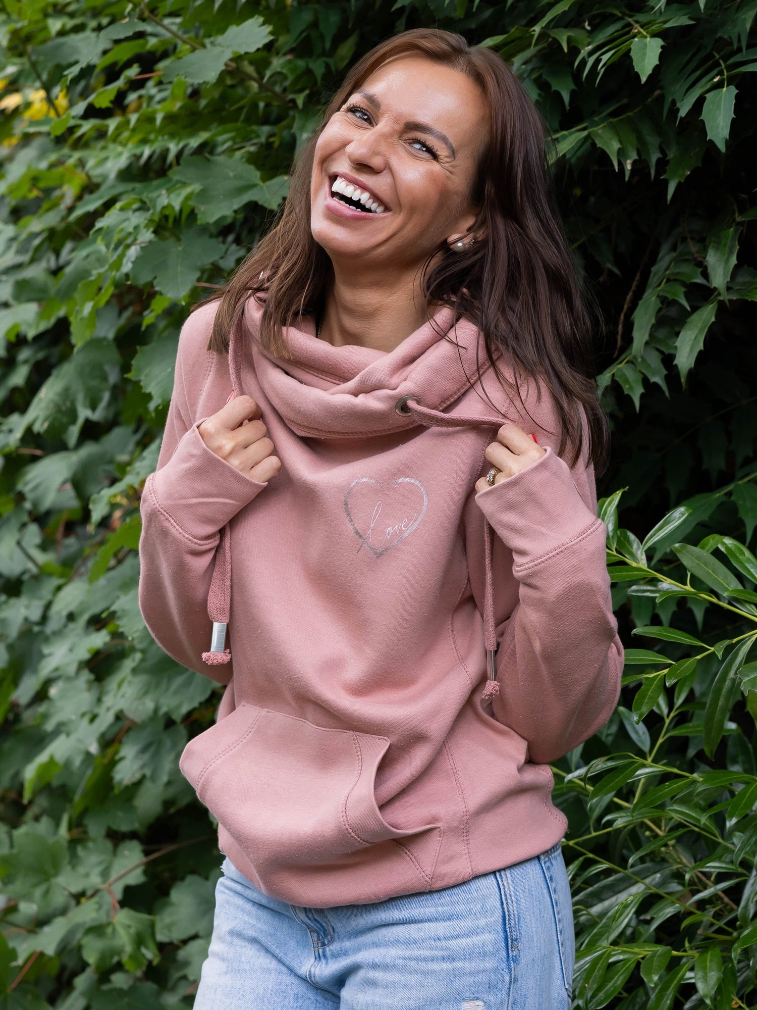 Model smiling outdoors in nature wears womens Cowl Neck Hoodie - Dusty Pink ladies hoodie with rose gold metallic love heart motif. Long sleeves and thumb loops to cuffs. Front kangaroo pouch pocket. Cross neck sweatshirt with hood