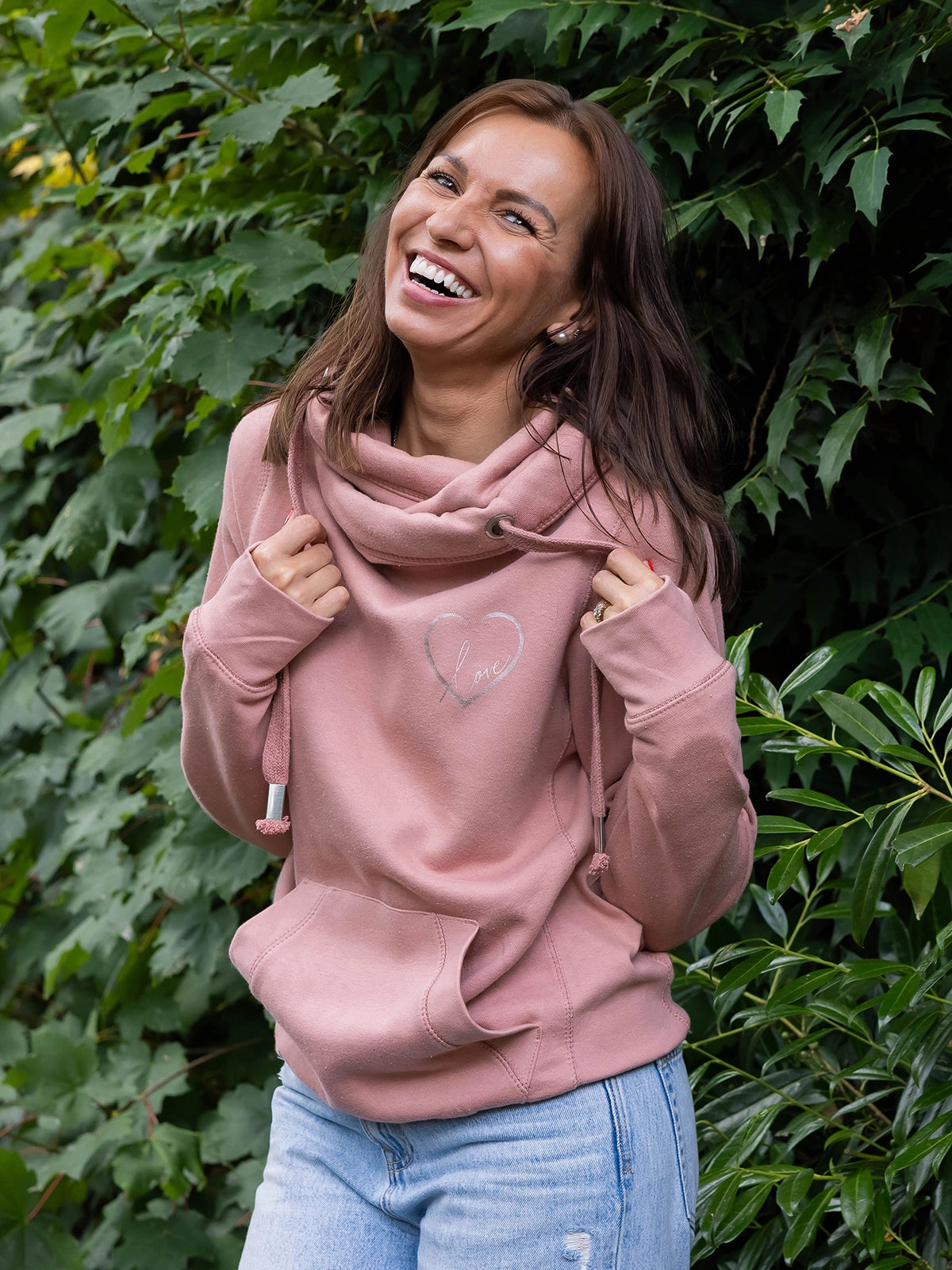 Model smiling outdoors in nature wears womens Cowl Neck Hoodie - Dusty Pink ladies hoodie with rose gold metallic love heart motif. Long sleeves and thumb loops to cuffs. Front kangaroo pouch pocket. Cross neck sweatshirt with hood