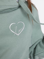 Womens Cowl Neck Hoodie - Close up of Dusty green hoodie with silver metallic love heart outline motif with the word love in silver script inside. Long sleeves Cross neck sweatshirt with hood and thick fabric drawstrings