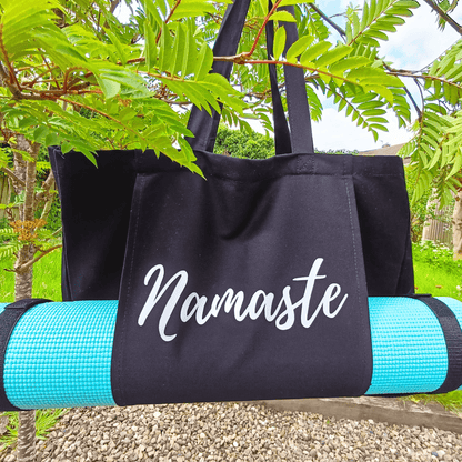 Black Yoga Mat tote bag with Silver script style writing Namaste across a front pouch pocket which holds a turqouise yoga mat length ways through the pouch. Mat is rolled and held you mat straps. Two shoulder straps. Photo is taken outdoors with tree leaves partially across the Large black tote bag hanging from a branch