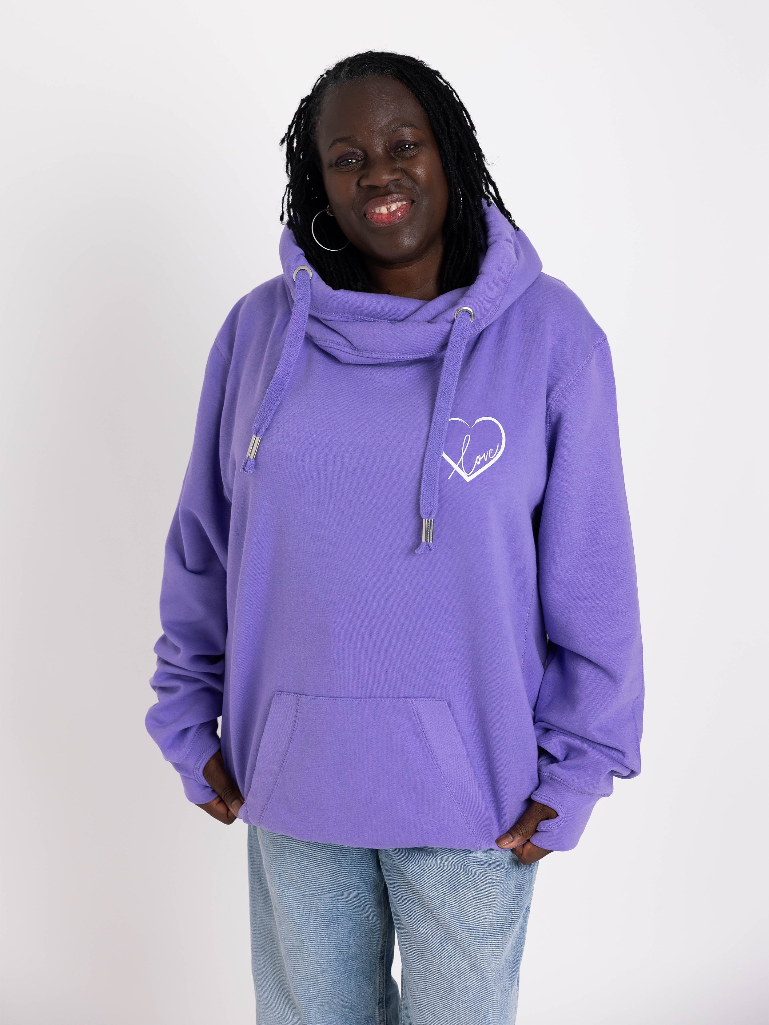 Woman wears a purple hoodie with a heart design on a plain background. lavender womens hoodie Ladies cowl neck fashion hoodie. Cotton Polyester blend relaxed fit unisex oversized style. White heart motif to upper left of the chest with word Love in script font nestled within the heart.