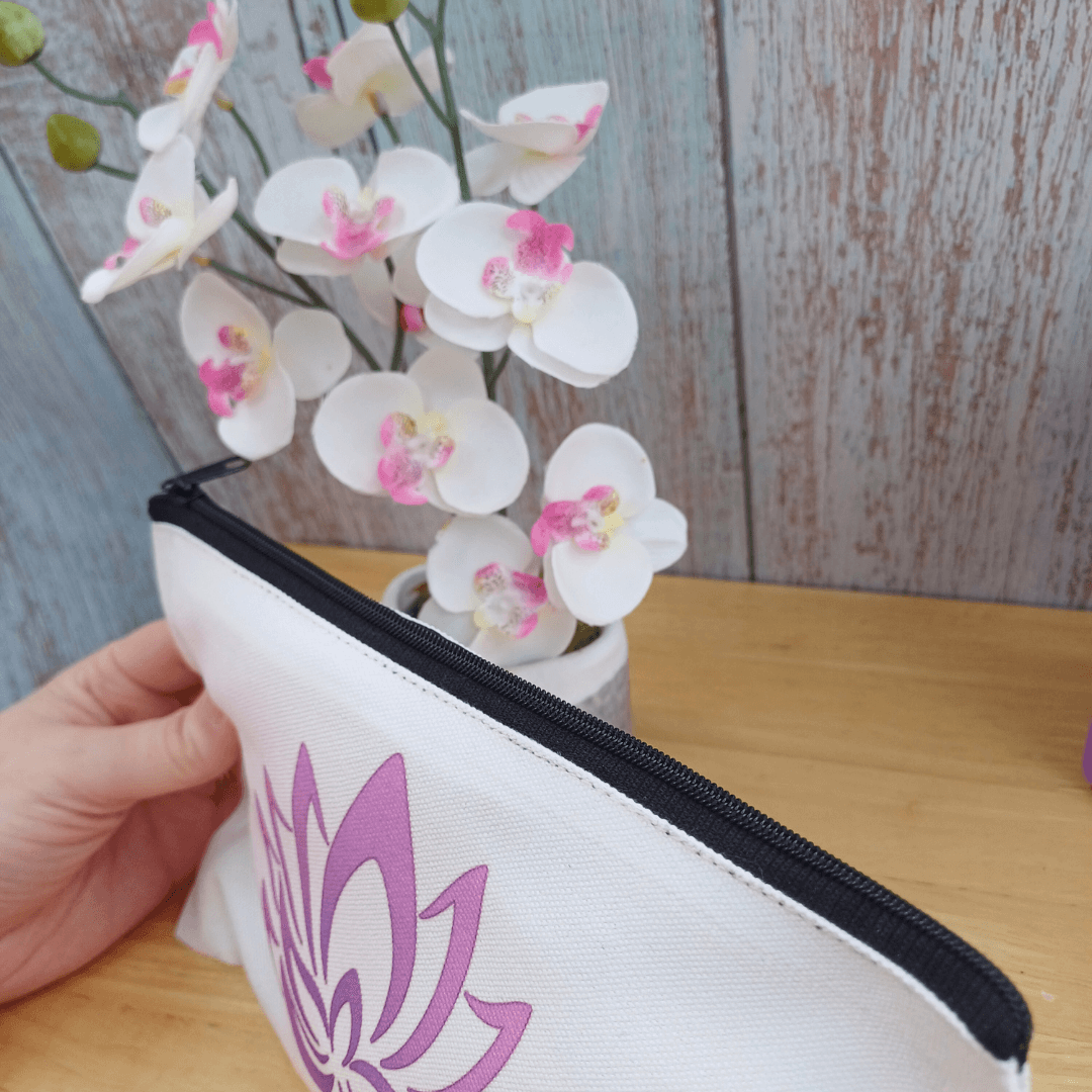 Yoga gift for her. Small personalised yoga theme make up bag. White with black zipped opening. Purple lotus flower design and name in purple below. White orchid and purple yoga block in background. Close up of black fabric zipper opening, fastened