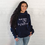 Womens Navy blue hoodie with cross cowl neck. Thumb holes to sleeves. Design is of four light grey butteflies in flight and wording Blue Skies and Butterflies to the front. Unisex relaxed fit hoodie. Light purple wording with light grey back shadow. Long sleeves and straps from cowl neck with metal toggles on the end. Worn by caucasian woman with long dark brown hair smiling 