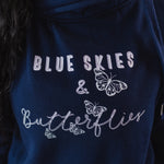 Close up of design on Womens Navy blue cross cowl neck hoodie. Four light grey butterflies and slogan Blue Skies and Butterflies to front. Unisex oversized fit. Light purple font with light grey back shadow. Long sleeves, metal toggle to drawstring