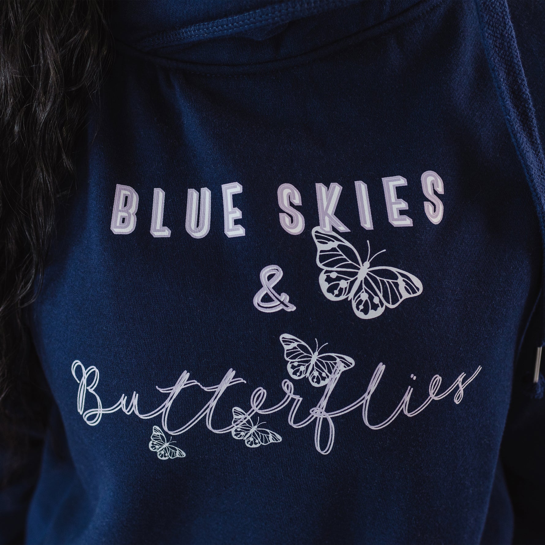 Close up of design on Womens Navy blue cross cowl neck hoodie. Four light grey butterflies and slogan Blue Skies and Butterflies to front. Unisex oversized fit. Light purple font with light grey back shadow. Long sleeves, metal toggle to drawstring