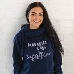 Womens Navy blue hoodie with cross cowl neck. Thumb holes to sleeves. Design is of four light grey butteflies in flight and wording Blue Skies and Butterflies to the front. Unisex relaxed fit hoodie. Light purple wording with light grey back shadow. Long sleeves and straps from cowl neck with metal toggles on the end. Worn by caucasian woman with long dark brown hair smiling 