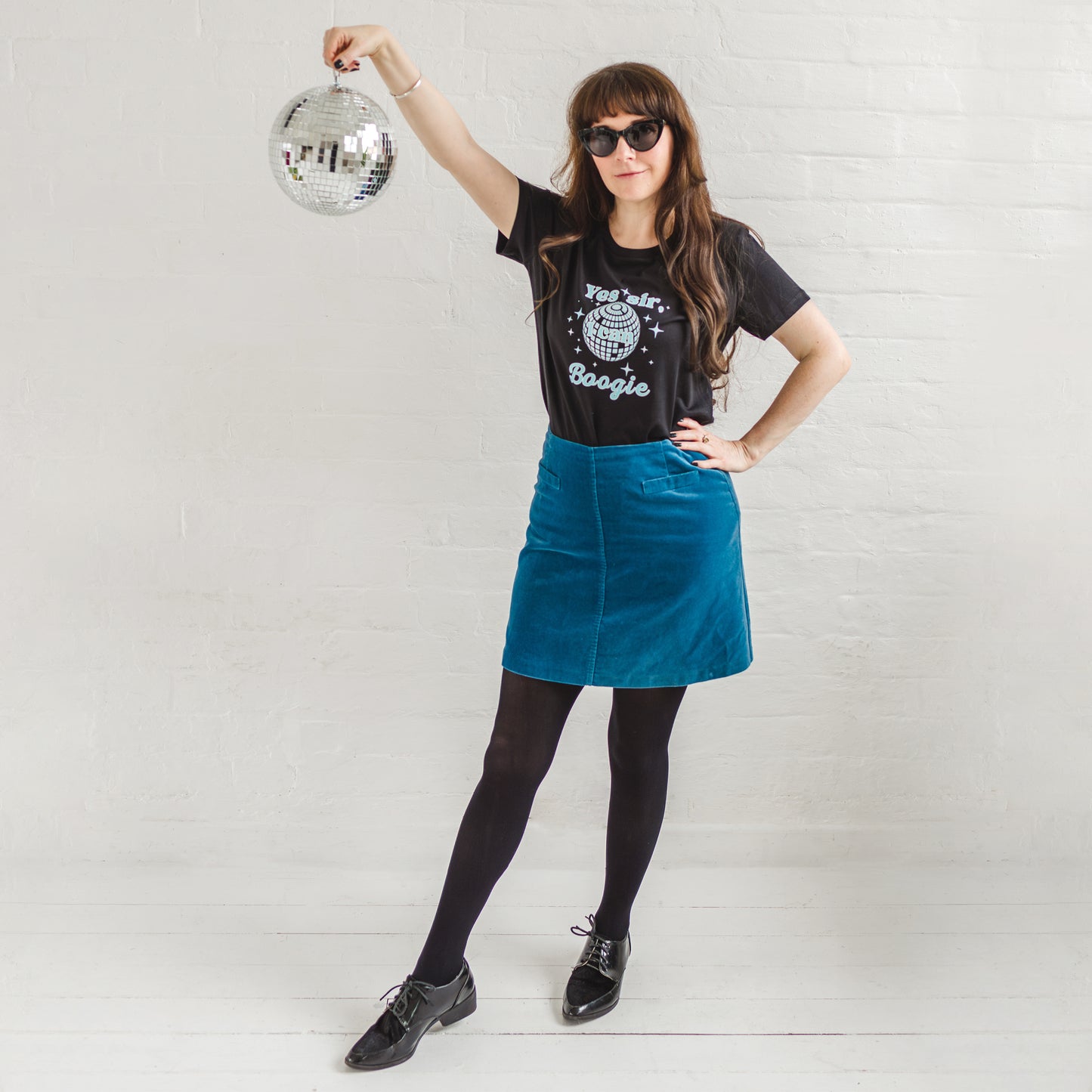 Black graphic t-shirt with mirror ball design.  Woman wearing sunglass smiling holding a silver disco ball, worn with a velvet A line mid blue skirt, black tights and black brogues. Fitted short sleeve tshirt with  round neck. 70s disco music inspired ladies slogan tshirt with disco ball design to background of top and sparkles surround. Slogan text in foreground reads Yes sir I can Boogie in light blue font. Organic cotton casual fitted Tee