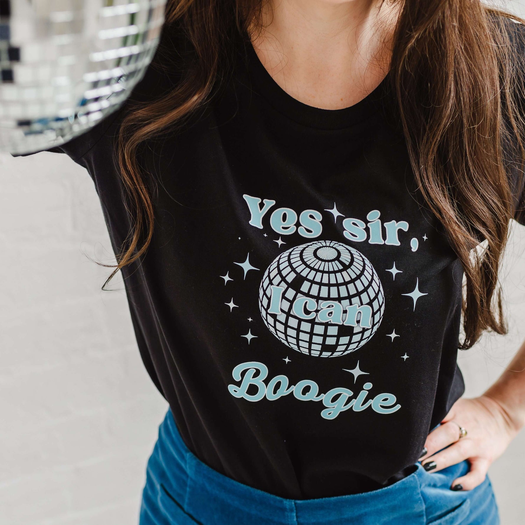 Close up of Ladies Black graphic t-shirt with mirror ball design. Woman holding a silver disco ball, worn with a velvet A line mid blue skirt. Fitted short sleeve tshirt with round neck. 70s disco music inspired ladies slogan tshirt with disco ball design to background of top and sparkles surround. Slogan text in foreground reads Yes sir I can Boogie in light blue font. Organic cotton casual fitted Tee