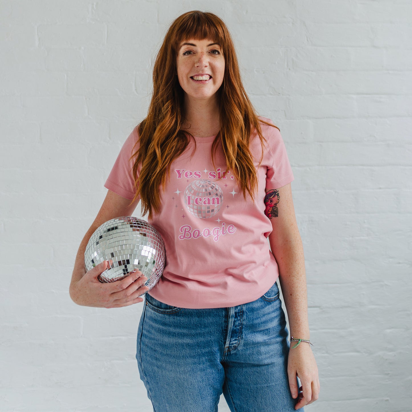 Ladies pink disco ball fitted t-shirt. Woman holding a silver disco ball, wears blue jeans and dusky pink round neck fitted tshirt. 70s disco music inspired womens pink graphic tshirt with silver tint mirrorball and sparkles. Slogan reads Yes sir I can Boogie in pink font with white outline
