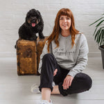 Woman sat on the floor smiling, wearing light grey sweatshirt without hood. Womens jumper unisex relaxed fit with crew neck and long sleeves with cuffs. Model wears black jeans. Slogan in black script font reads French phrase C'est Magnifique, Behind her there is a wooden weathered chest and a black cockapoo dog sitting on it happy and looking to camera with it's tongue out