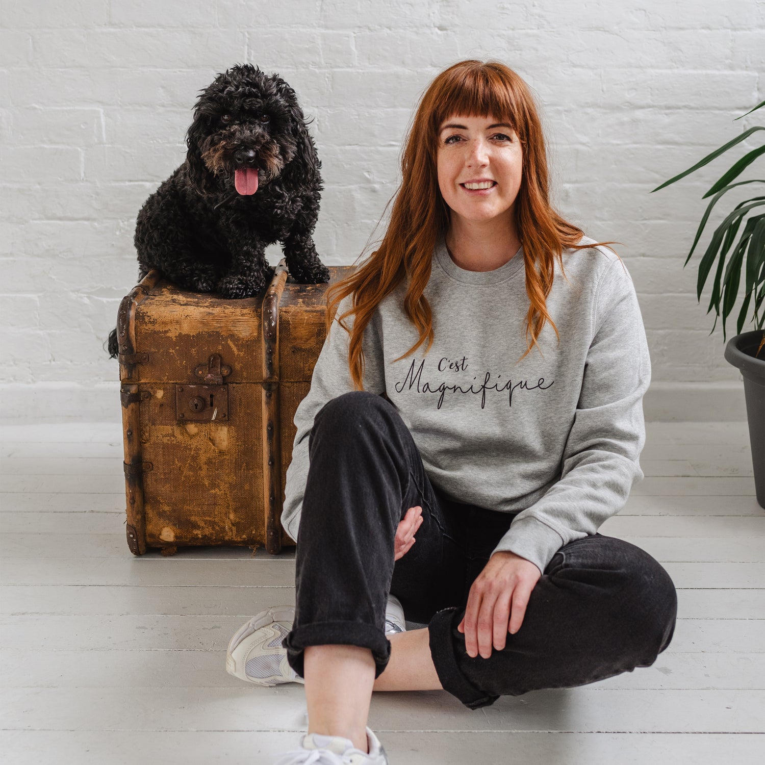 Woman sat on the floor smiling, wearing light grey sweatshirt without hood. Womens jumper unisex relaxed fit with crew neck and long sleeves with cuffs. Model wears black jeans. Slogan in black script font reads French phrase C'est Magnifique, Behind her there is a wooden weathered chest and a black cockapoo dog sitting on it happy and looking to camera with it's tongue out