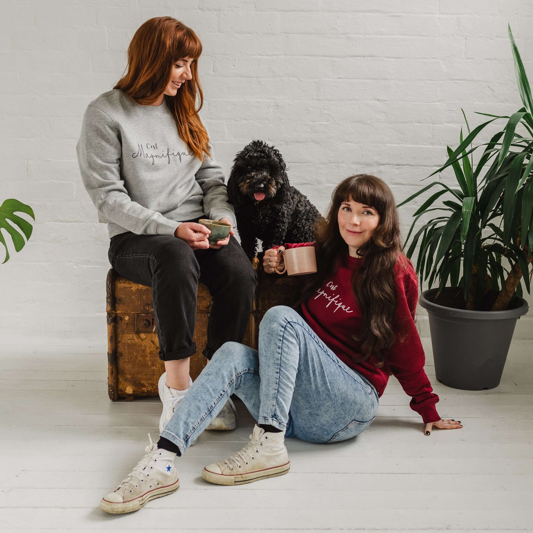 Two women, woman wearing burgundy red sweatshirt is sat on the floor, jumper is unisex relaxed fit with crew neck and long sleeves with cuffs. Model wears light blue acid wash jeans. Slogan in white script font reads French phrase C'est Magnifique, Behind her there is a wooden weathered chest and a black cockapoo dog sitting on it looking forward with it's tongue out. A woman wearing a matching design light grey ladies sweatshirt sits on the chest looking down at the dog smiling. Both hold cups of coffee.