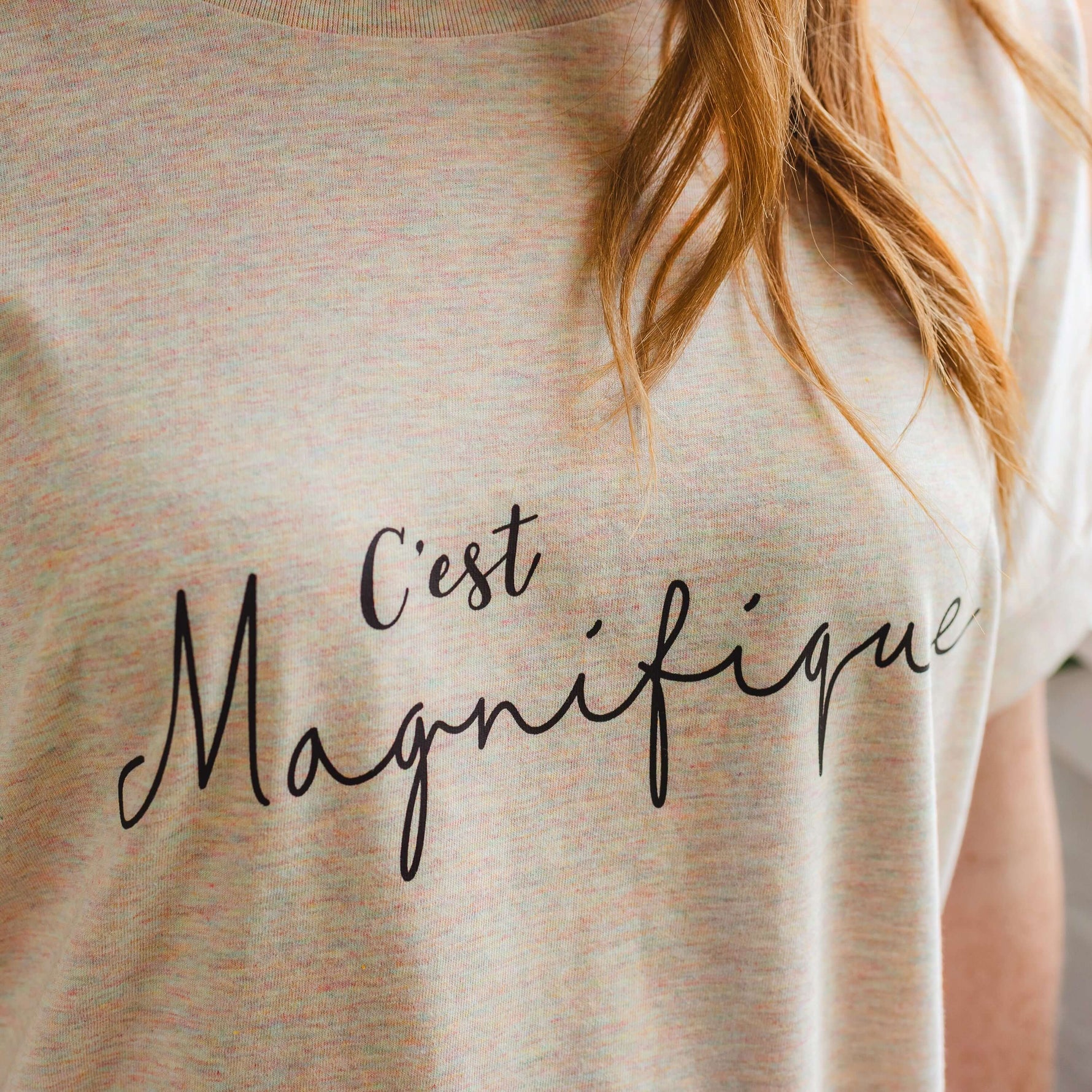 Close up image of womens fashion t shirt in light beige / cream with subtle rainbow multicolour flecks. Black script slogan text across chest reads C'est Magnifique