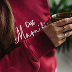 Close up of womens burgundy red trendy sweatshirt with white slogan script wording C'est Magnifique. Woman is holding a coffee cup 