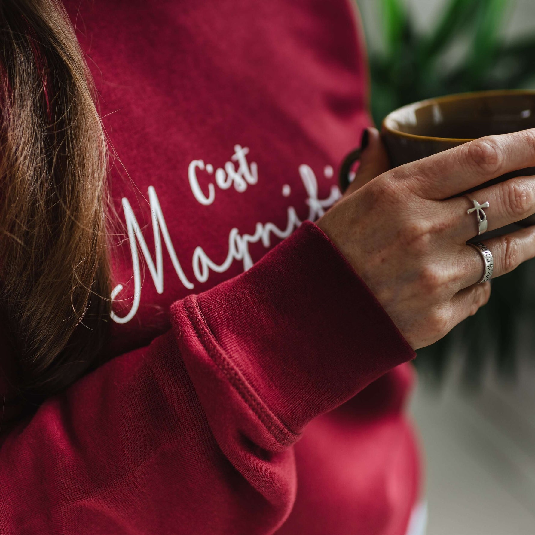 Close up of womens burgundy red trendy sweatshirt with white slogan script wording C'est Magnifique. Woman is holding a coffee cup 