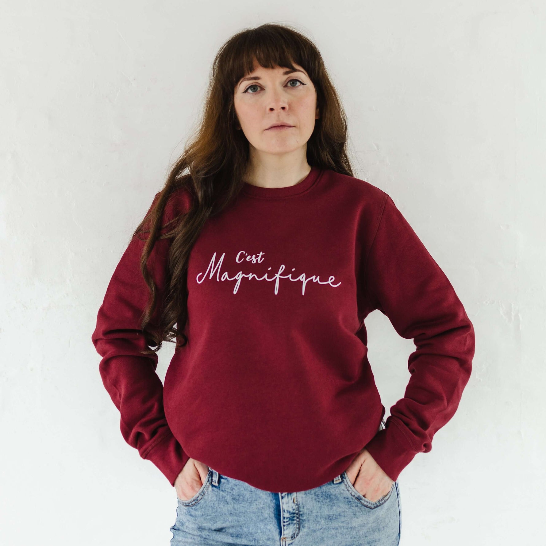 Caucasian woman with long brown hair wearing blue acid wash jeans and a burgundy red relaxed fit sweatshirt jumper with crew round neck and long sleeves with cuffs. 