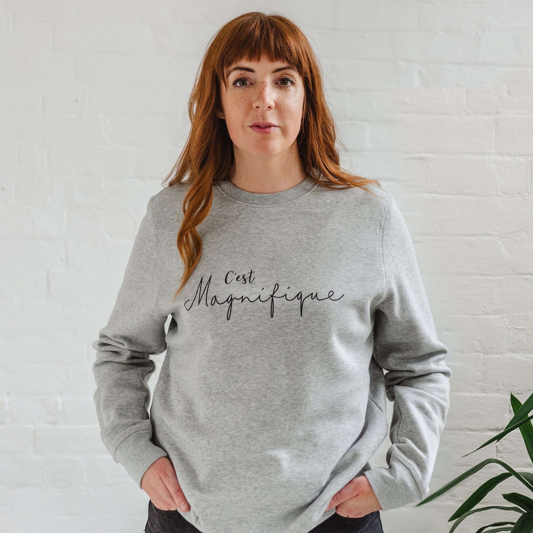 Woman wearing light grey sweatshirt without hood. Womens jumper unisex relaxed fit with crew neck and long sleeves with cuffs. Model wears black jeans. Slogan in black script font reads French phrase C'est Magnifique 