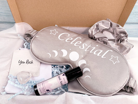 Letterbox relaxation gift set. Aromatherapy gift set with white tissue paper gift wrap. Silver satin sleep mask with stars and white moon cycles and wording Celestial, satin hair scrunchie. Essential oil Anti anxiety roll-on. Clear quartz healing crystal, card insert 'You Rock'
