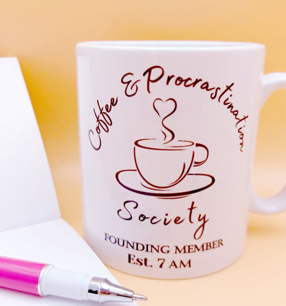 Coffee lover ceramic mug gift. White coffee mug with cup and saucer design. Black outline with steam up from cup shaped into a heart. Writing above design reads Coffee & Procrastination. Below design reads ...Society FOUNDING MEMBER Est. 7am Blank notepad and pen to left of the 10oz mug