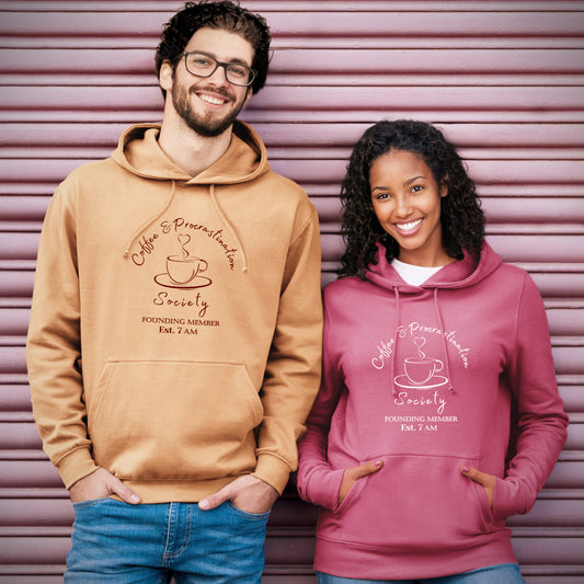Man and woman matching unisex relaxed fit hoodie. Man's caramel with dark brown design. Woman's plum with white design. Coffee lover theme coffee cup and saucer and heart shaped steam. Slogan Coffee & Procrastination Society FOUNDING MEMBER Est. 7am Hood and fabric drawstrings.