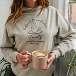 Woman with auburn long hair standing near a window holding a mug of coffee wears a Ladies coffee themed oversized fit hoodie. Stone grey hoodie top with coffee cup and saucer design. Black outline with steam from cup shaped into a heart. Writing above reads Coffee & Procrastination. Below reads Society FOUNDING MEMBER Est. 7am.