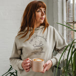 Woman with auburn long hair standing looking out a window holding a mug of coffee wears a Ladies coffee themed oversized fit hoodie. Stone grey hoodie top with coffee cup and saucer design. Black outline with steam from cup shaped into a heart. Writing above reads Coffee & Procrastination. Below reads Society FOUNDING MEMBER Est. 7am.