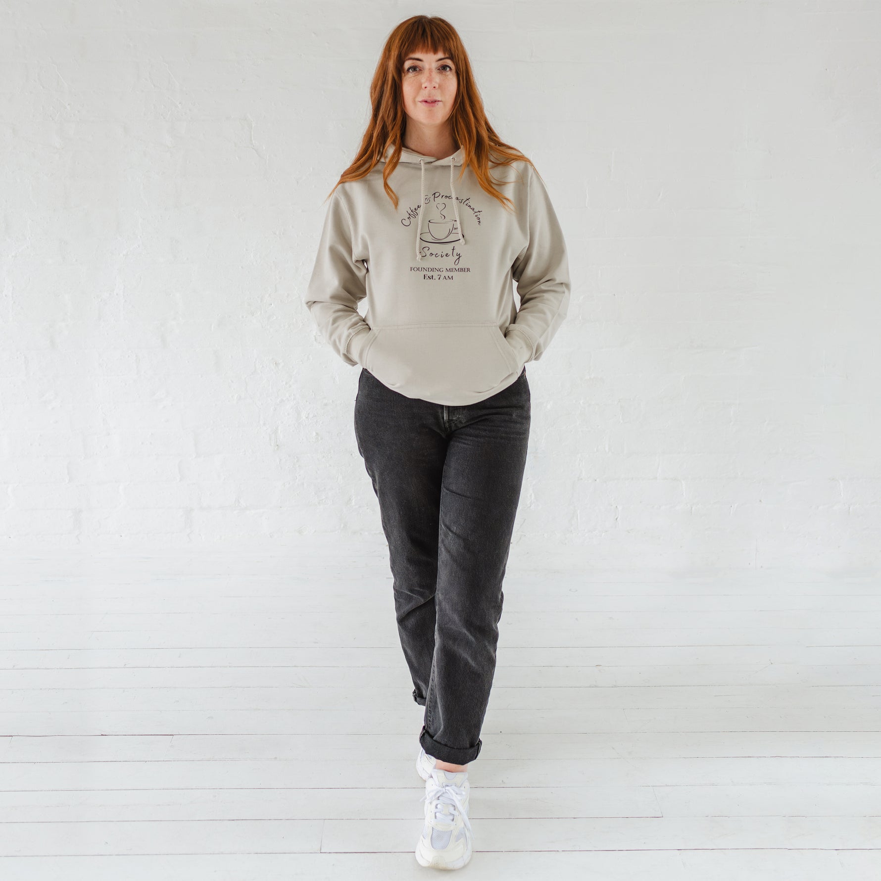 Woman with auburn long hair wears a womens coffee themed oversized fit hoodie. Stone grey hoodie top with coffee cup and saucer design. Black outline with steam from cup shaped into a heart. Writing above reads Coffee & Procrastination. Below reads Society FOUNDING MEMBER Est. 7am. Worn with black straight leg jeans and white trainers 