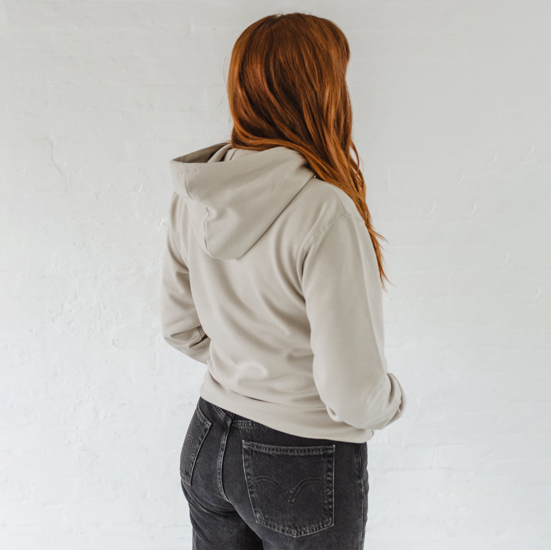 Woman with auburn long hair standing with her back turned to show hood  wears a Ladies coffee themed oversized fit hoodie. Stone grey hoodie top with coffee cup and saucer design. Black outline with steam from cup shaped into a heart. Writing above reads Coffee & Procrastination. Below reads Society FOUNDING MEMBER Est. 7am. Worn with black straight leg jeans