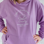 Close up of lavender purple coffee lovers hoodie. Woman with blonde long hair wears a Ladies coffee themed oversized fit hoodie. Lilac / Lavender unisex long sleeve hoodie top with coffee cup and saucer design. White outline with steam from cup shaped into a heart. Writing above reads Coffee & Procrastination. Below reads Society FOUNDING MEMBER Est. 7am.