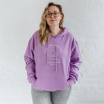 Woman with blonde long hair standing wears a womens coffee themed oversized fit hoodie. Lavender purple hoodie top with coffee cup and saucer design. White outline with steam from cup shaped into a heart. Writing above reads Coffee & Procrastination. Below reads Society FOUNDING MEMBER Est. 7am. She pairs it with grey skinny jeans