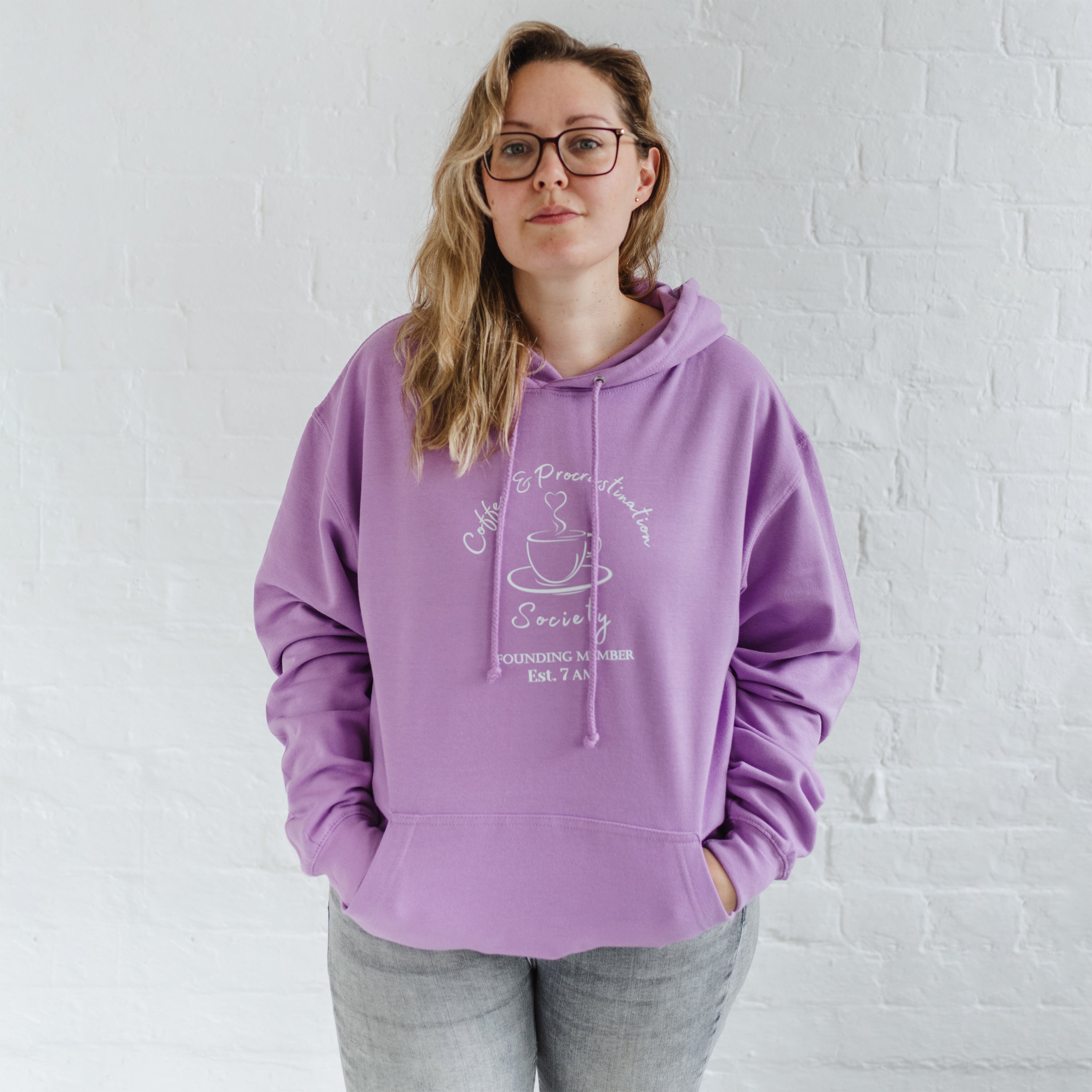 Woman with blonde long hair standing wears a womens coffee themed oversized fit hoodie. Lavender purple hoodie top with coffee cup and saucer design. White outline with steam from cup shaped into a heart. Writing above reads Coffee & Procrastination. Below reads Society FOUNDING MEMBER Est. 7am. She pairs it with grey skinny jeans