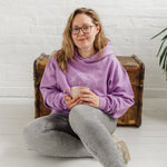 Woman with blonde long hair seated with a mug of coffee wears a Ladies coffee themed oversized fit hoodie. Lavender purple hoodie top with coffee cup and saucer design. White outline with steam from cup shaped into a heart. Writing above reads Coffee & Procrastination. Below reads Society FOUNDING MEMBER Est. 7am. She pairs it with grey skinny jeans and trainers. 