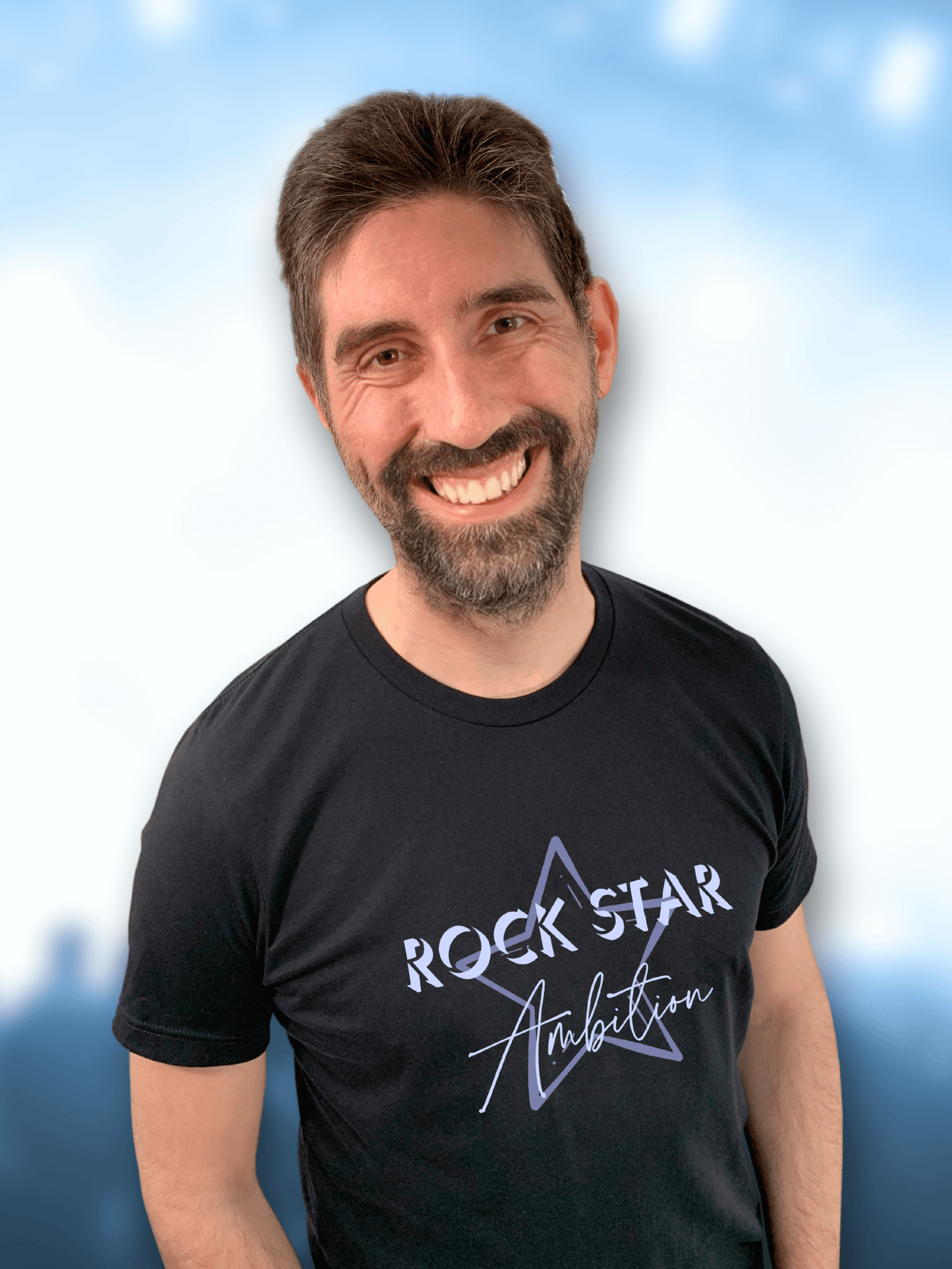 Concert background with Man wearing mens black short sleeve graphic tshirt. Crew neck casual wear. Band Tee Design is mid purple star outline behind wording ROCK STAR in upper case shadowed light grey/purple text, then Ambition below in script. Rock music themed unisex top