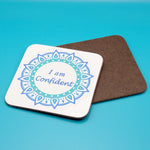 White positive affirmation coaster with blue mandala design. Daily affirmation reads I am Confident. Coaster underneath shows wood backing